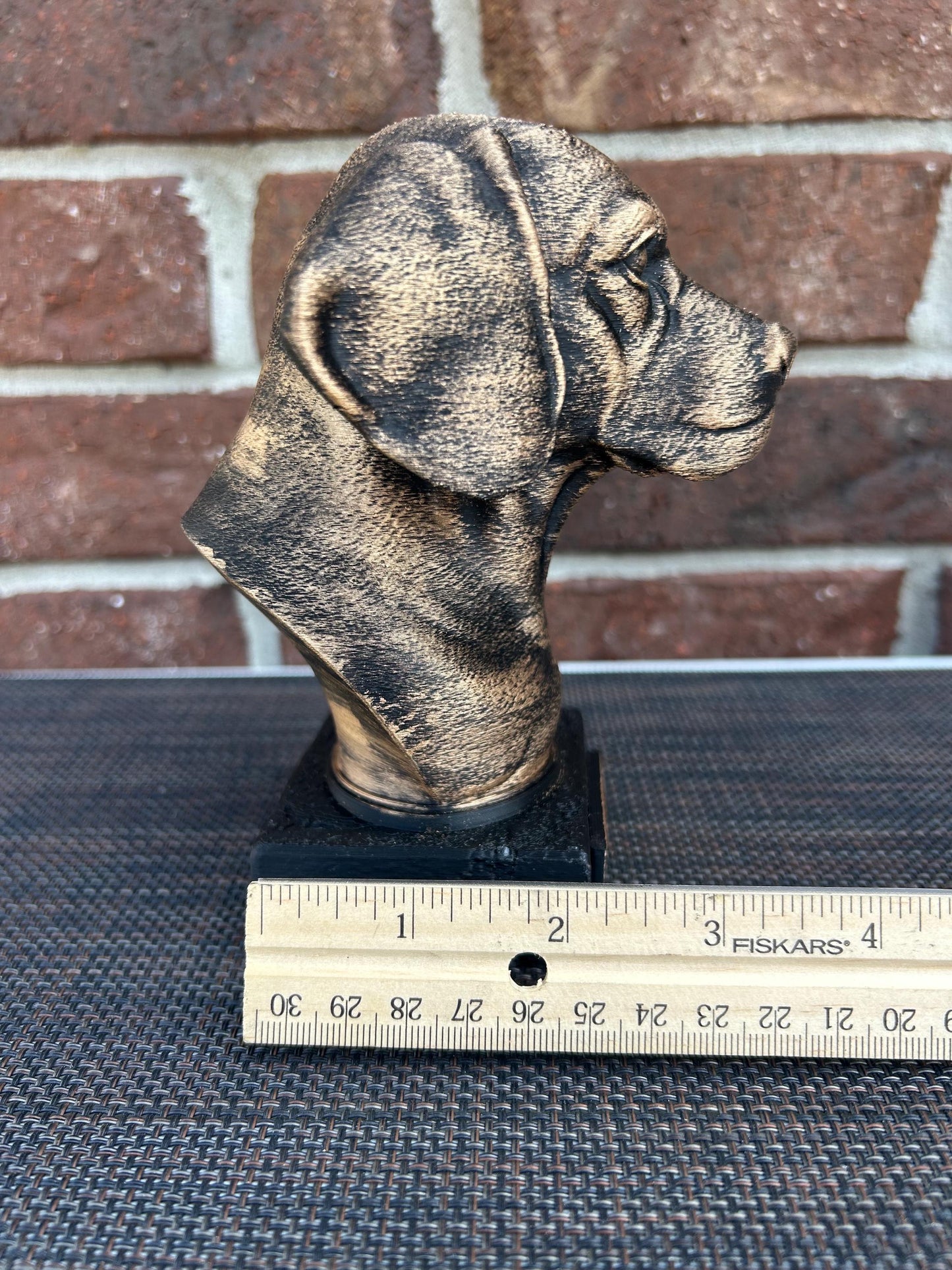 The dog bust is a small, bronze-like sculpture on a rectangular base. Viewing the right profile, a floppy ear hangs down. The fur is detailed with fine, wavy lines around its neck and chest, giving a thick, textured feel. The surface is smooth but weathered, with a faint patina adding a rough texture in spots. The statue stands about 6 inches tall, capturing the essence of a beagle with a calm, attentive expression.