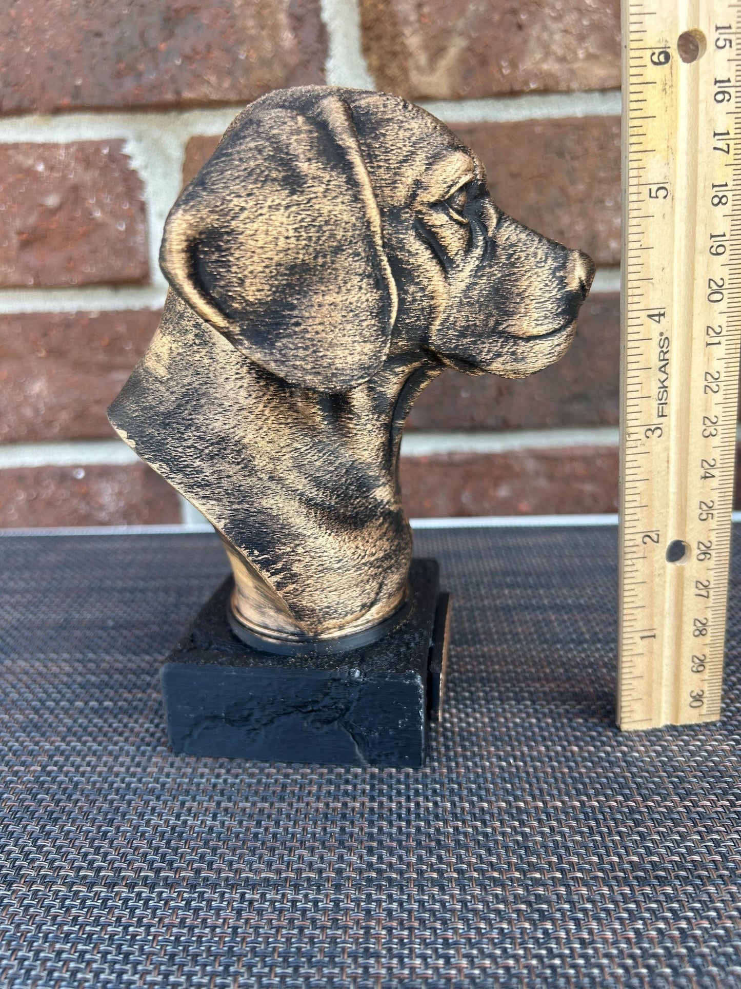 The dog bust is a small, bronze-like sculpture on a rectangular base. Viewing the left profile, a floppy ear hangs down. The fur is detailed with fine, wavy lines around its neck and chest, giving a thick, textured feel. The surface is smooth but weathered, with a faint patina adding a rough texture in spots. The statue stands about 6 inches tall, capturing the essence of a beagle with a calm, attentive expression.