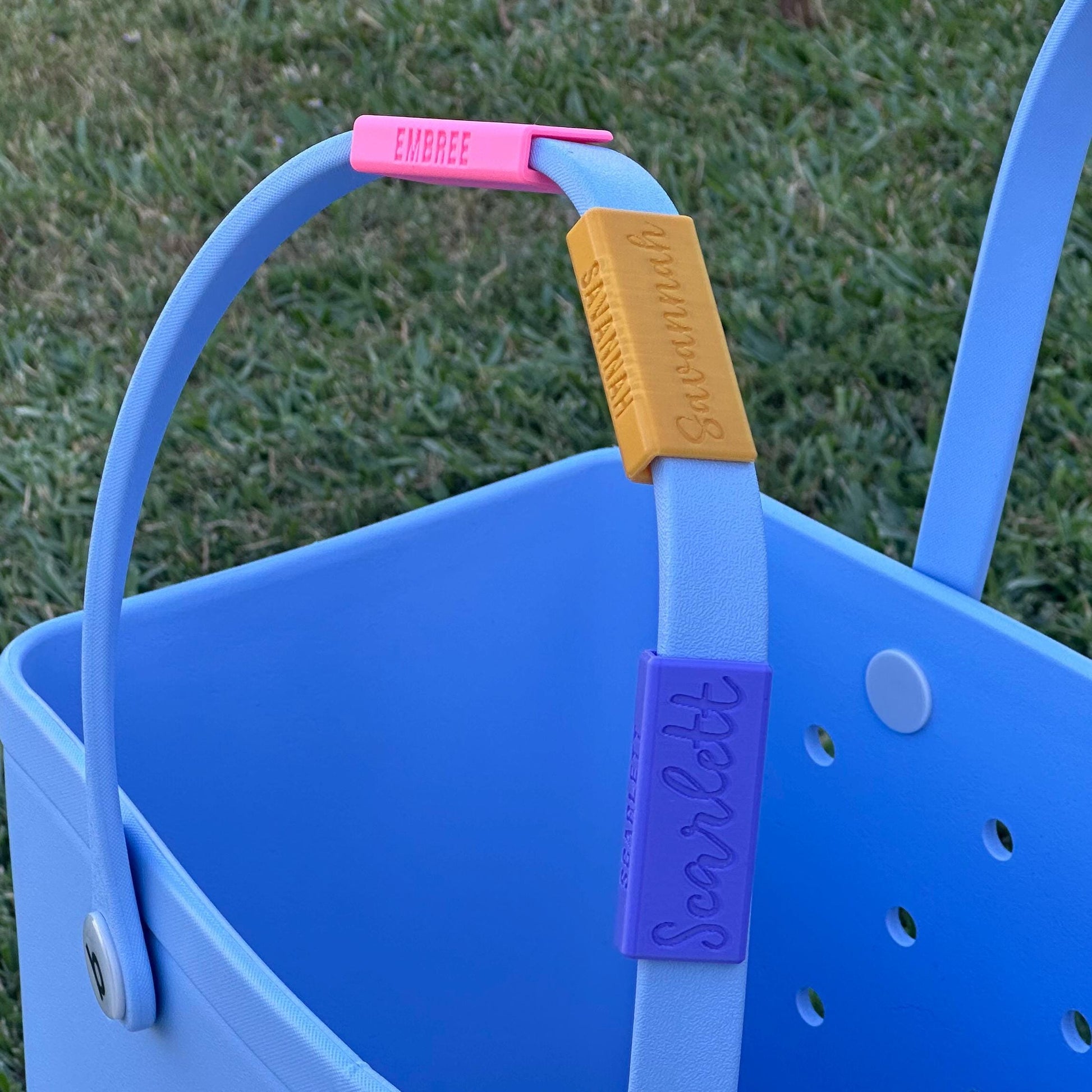 Multiple custom name clips, in pink, golden yellow, and blue on a bogg bag handle