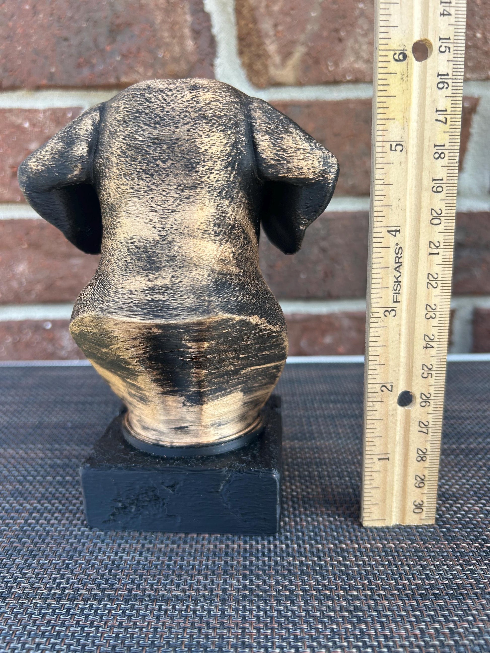 The dog bust is a small, bronze-like sculpture on a rectangular base. Viewing the back of the head, floppy ears hanging down. The fur is detailed with fine, wavy lines around its neck and chest, giving a thick, textured feel. The surface is smooth but weathered, with a faint patina adding a rough texture in spots. The statue stands about 6 inches tall, capturing the essence of a beagle with a calm, attentive expression.