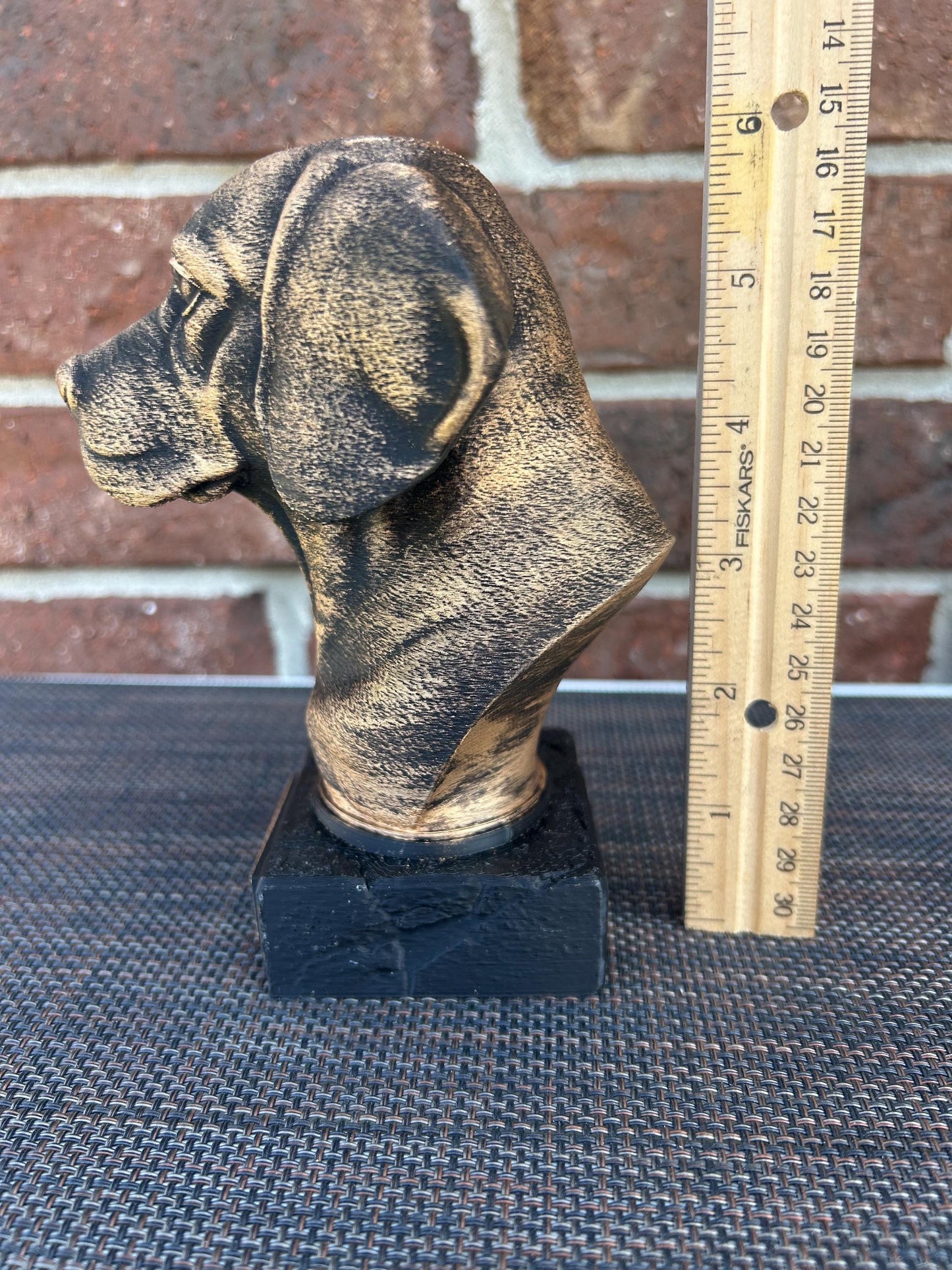 The dog bust is a small, bronze-like sculpture on a rectangular base. Viewing the left profile, a floppy ear hangs down. The fur is detailed with fine, wavy lines around its neck and chest, giving a thick, textured feel. The surface is smooth but weathered, with a faint patina adding a rough texture in spots. The statue stands about 6 inches tall, capturing the essence of a beagle with a calm, attentive expression.