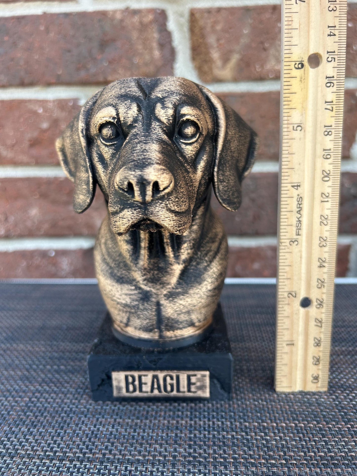 The dog bust is a small, bronze-like sculpture on a rectangular base, labeled &quot;BEAGLE&quot; on a plaque. Its head looks straight forward, with floppy ears hanging down. The fur is detailed with fine, wavy lines around its neck and chest, giving a thick, textured feel. The surface is smooth but weathered, with a faint patina adding a rough texture in spots. The statue stands about 6 inches tall, capturing the essence of a beagle with a calm, attentive expression.