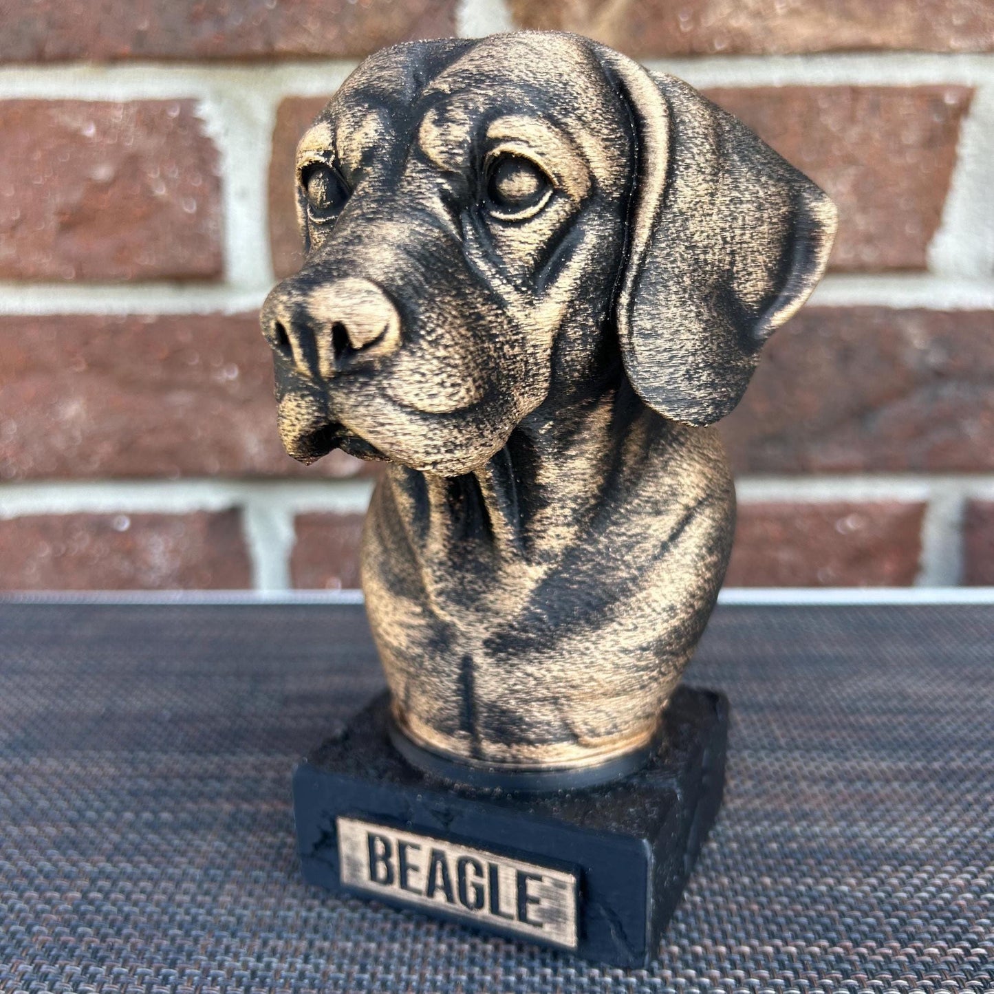 The dog bust is a small, bronze-like sculpture on a rectangular base, labeled &quot;BEAGLE&quot; on a plaque. Its head tilts slightly to the left, with floppy ears hanging down. The fur is detailed with fine, wavy lines around its neck and chest, giving a thick, textured feel. The surface is smooth but weathered, with a faint patina adding a rough texture in spots. The statue stands about 6 inches tall, capturing the essence of a beagle with a calm, attentive expression.