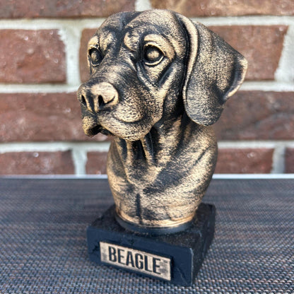 The dog bust is a small, bronze-like sculpture on a rectangular base, labeled &quot;BEAGLE&quot; on a plaque. Its head tilts slightly to the left, with floppy ears hanging down. The fur is detailed with fine, wavy lines around its neck and chest, giving a thick, textured feel. The surface is smooth but weathered, with a faint patina adding a rough texture in spots. The statue stands about 6 inches tall, capturing the essence of a beagle with a calm, attentive expression.