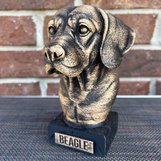 The dog bust is a small, bronze-like sculpture on a rectangular base, labeled &quot;BEAGLE&quot; on a plaque. Its head tilts slightly to the left, with floppy ears hanging down. The fur is detailed with fine, wavy lines around its neck and chest, giving a thick, textured feel. The surface is smooth but weathered, with a faint patina adding a rough texture in spots. The statue stands about 6 inches tall, capturing the essence of a beagle with a calm, attentive expression.